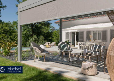 Una pérgola bioclimática adosada a una casa a modo de terraza. La pérgola, además de las lamas superiores, tiene toldos en los lados para tapar del sol la estancia completamente.